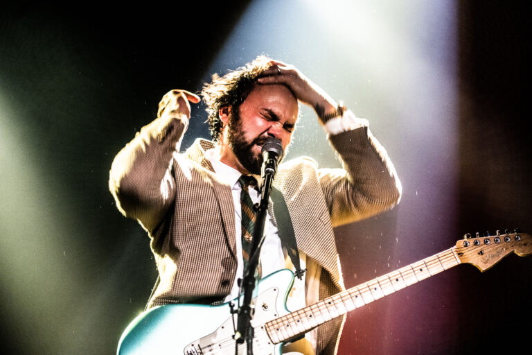 Een man met een gitaar zingt energiek op een podium, terwijl hij zijn hoofd vasthoudt.