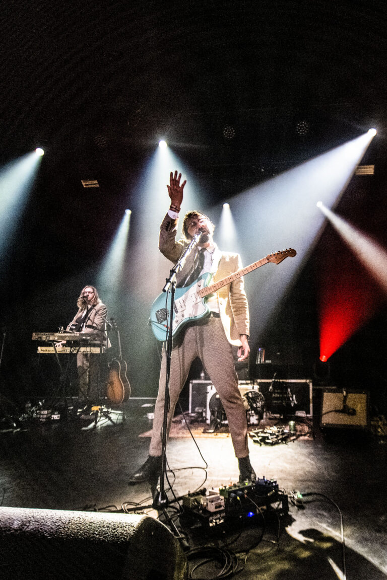 Een uitvo performer met een gitaar staat in de schijnwerpers, terwijl een tweede muzikant op een keyboard speelt.