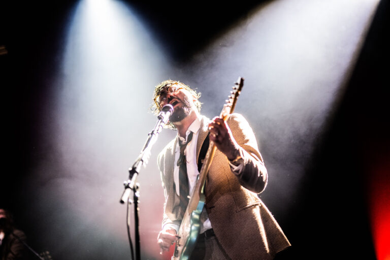 Een muzikant in een lichtgrijs pak staat op het podium en zingt in een microfoon, omgeven door sfeervolle verlichting.