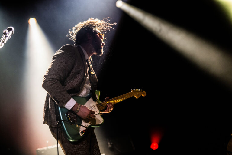 Een muzikant speelt gitaar op het podium, met dynamische verlichting en een levendige uitstraling.