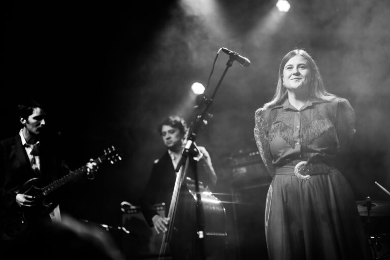 Een zwart-witfoto van drie muzikanten op het podium, met een microfoon en instrumenten, onder een sfeervolle verlichting.