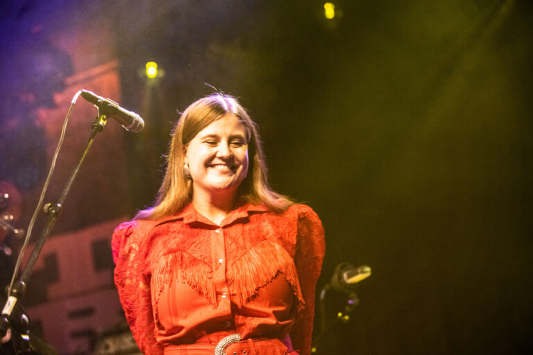 Een vrouw in een felrode outfit staat op het podium, met een microfoon in de buurt en een vrolijke uitstraling.