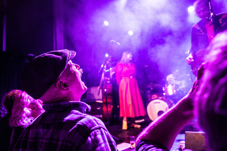 Een publiek kijkt op naar een muzikant in een opvallende rode outfit, omringd door paarse lichten en rook op het podium.