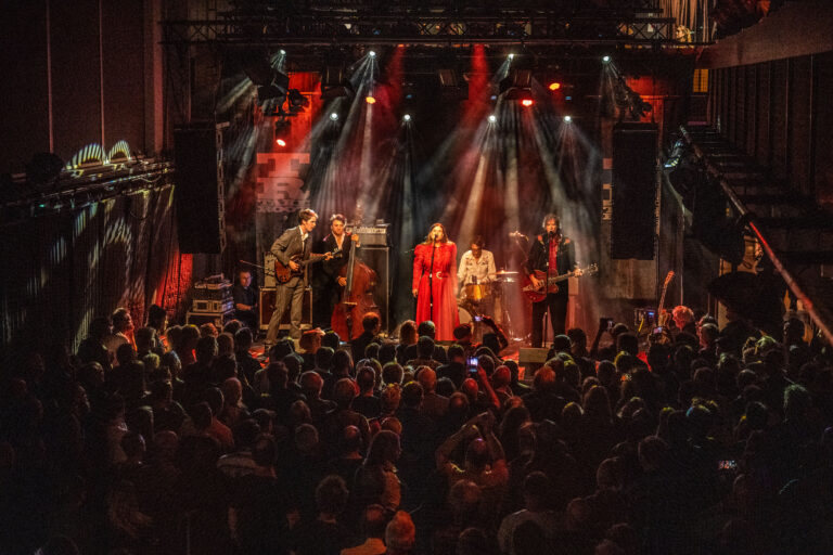 Een band treedt op voor een enthousiast publiek, omgeven door kleurrijke verlichting op het podium.