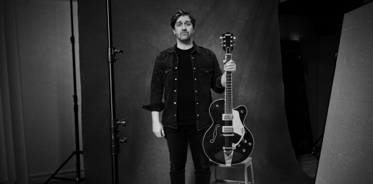 Een man staat met een zwarte gitaar naast een stoel voor een neutrale achtergrond.