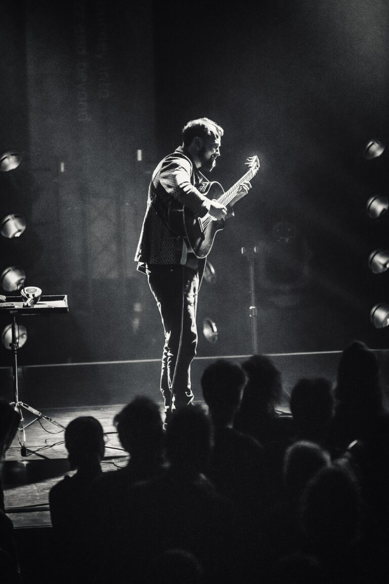 Een muzikant speelt gitaar op een podium, verlicht door spots, met een publiek ervoor.