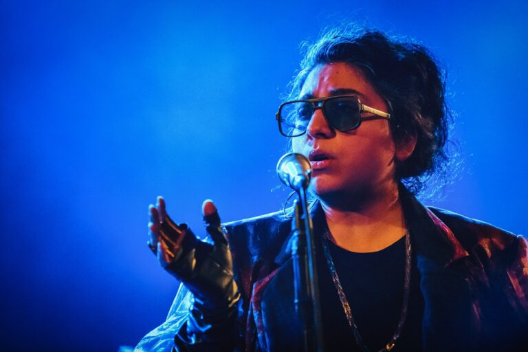 Een zanger in een zwart jasje en zonnebril, die zingt in een blauwe lichtshow.