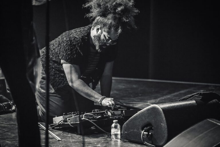 Een muzikant met een opvallende haardos werkt aan apparatuur op het podium.