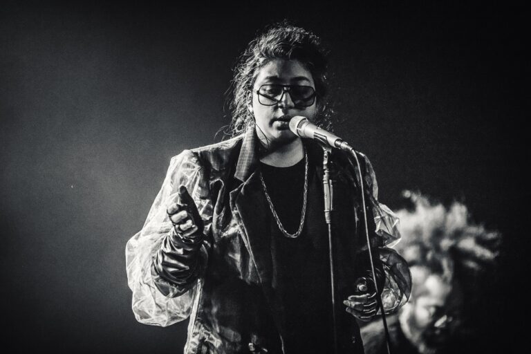 Een zangeres met een microphone in de hand, gekleed in een lichtgekleurde jas, staat op een podium met een dramatische lichtsetting.