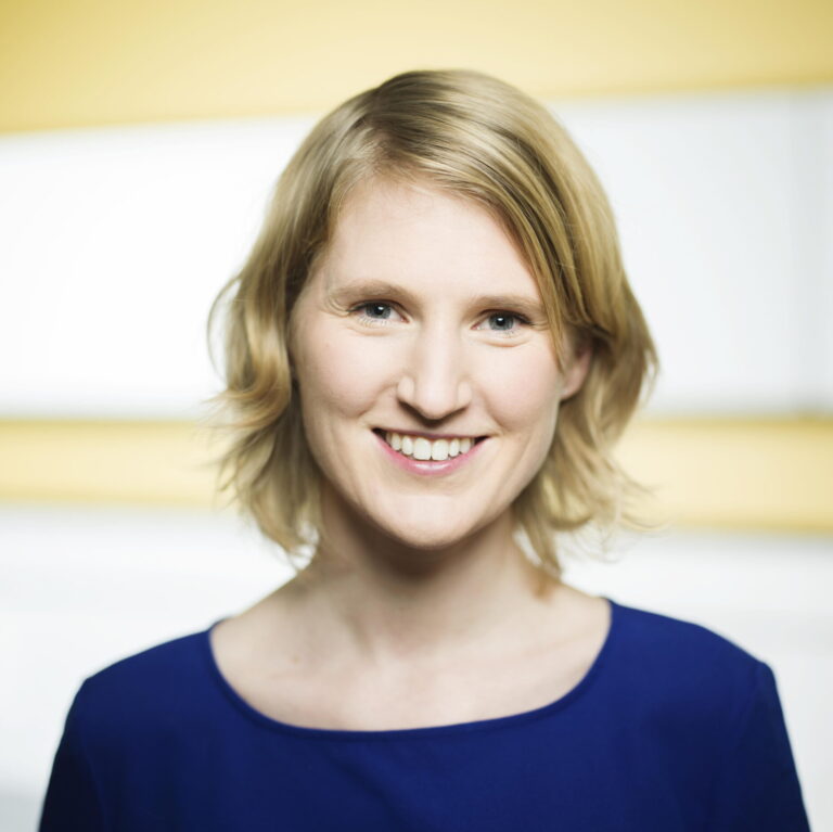 Portret van een glimlachende vrouw met kort, blond haar, gekleed in een blauwe top, tegen een lichte achtergrond.