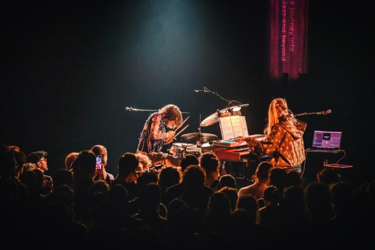 Een live optreden van twee muzikanten die enthousiast spelen voor een aanwezige menigte in een donkere concertzaal.