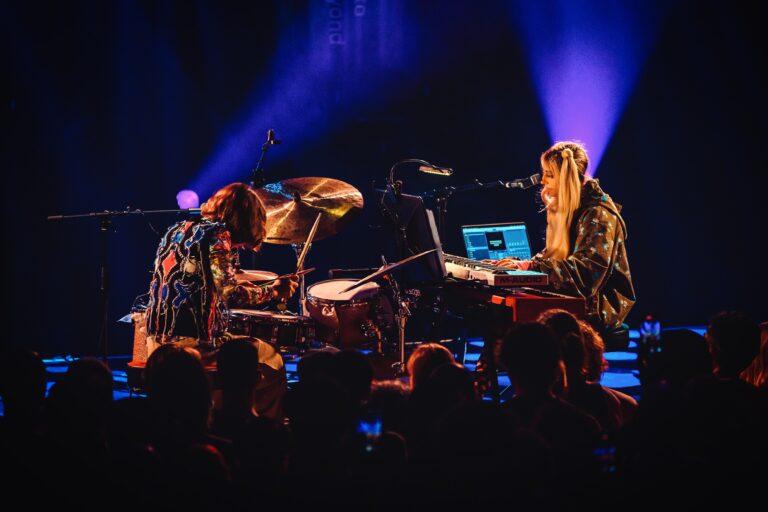 Twee muzikanten spelen live op een podium met kleurrijke verlichting.