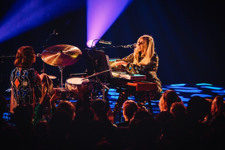 Een muzikant speelt piano en zingt op een podium, terwijl het publiek toekijkt in een sfeervolle verlichting.