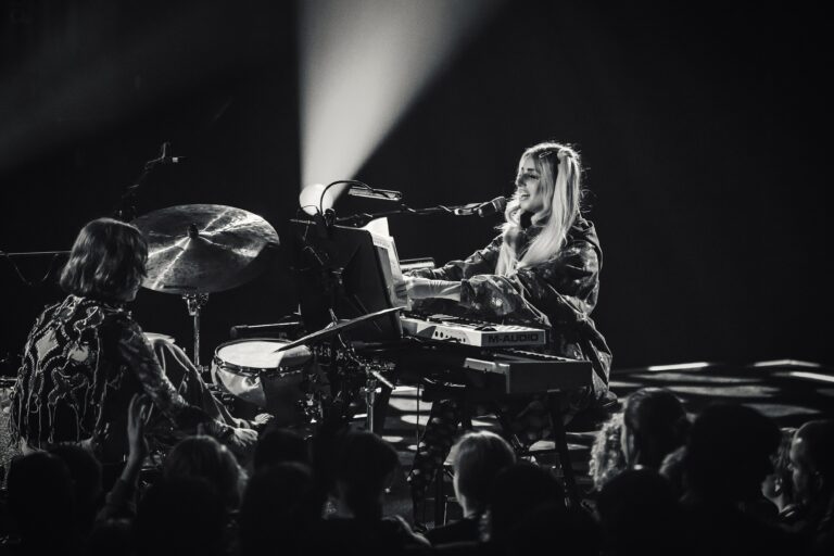 Een muzikante speelt op een keyboard en drumstel terwijl ze in schijnwerpers staat.