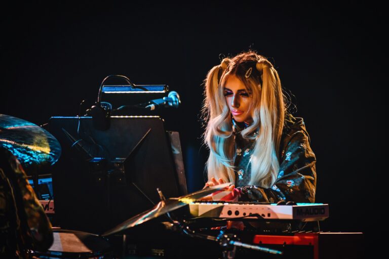 Een muzikante speelt op een keyboard met een microfoon en heeft lang, steil haar in een lichte tint.