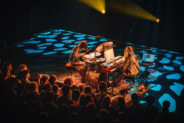 Twee muzikanten spelen op een podium voor een publiek, met kleurrijke lichteffecten op de vloer.