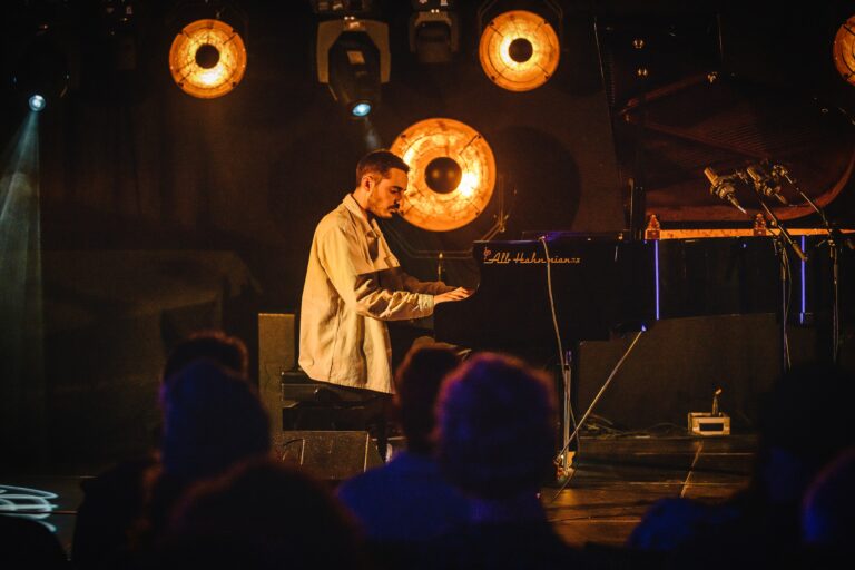 Een muzikant speelt piano op een podium, omgeven door zacht licht van vintage lampen en een aandachtig publiek.