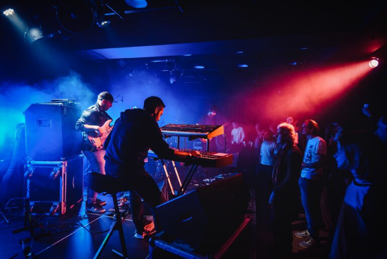 Een liveband speelt op het podium terwijl het publiek geniet van de muziek in een sfeervolle, kleurrijke lichte omgeving.