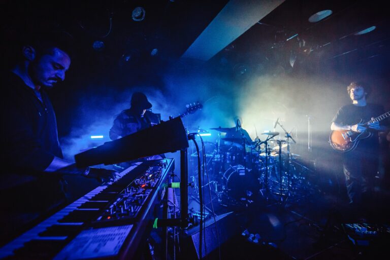 Een muzikant speelt op een keyboard met een drummer en kleurrijke lichteffecten op de achtergrond.