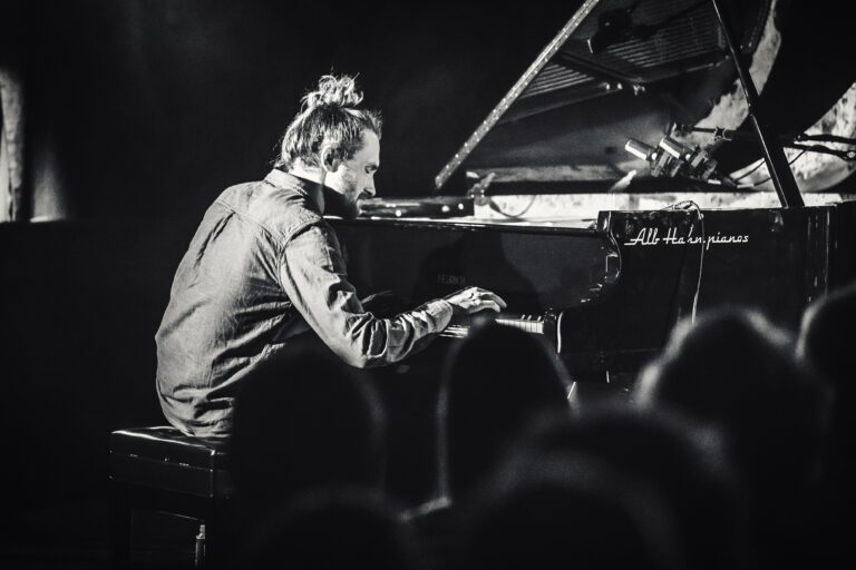 Een muzikant speelt piano op een podium, zichtbaar in een zwart-wit sfeer met een publiek op de achtergrond.