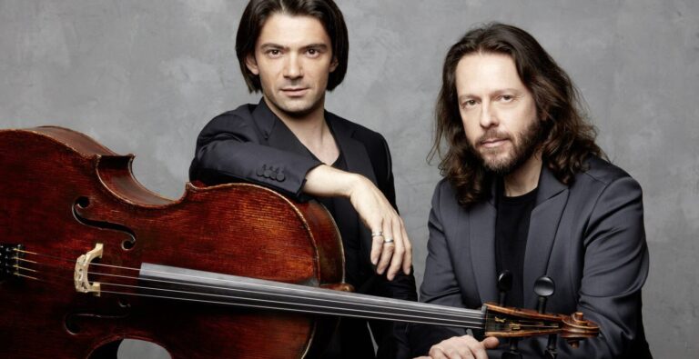 Twee mannen, een met een cello, poseren samen voor een neutrale achtergrond.