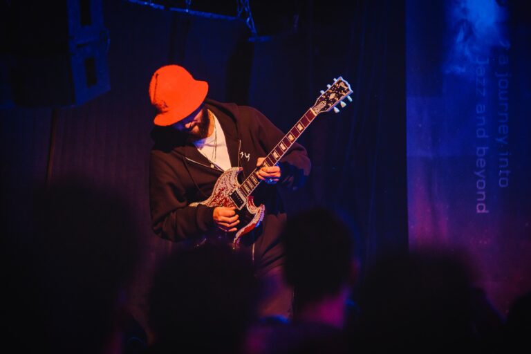 Een muzikant speelt gitaar op het podium, gekleed in een zwarte outfit en een oranje hoed.