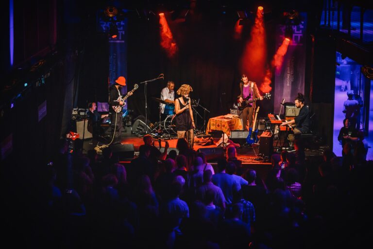 Een band speelt live op een podium met kleurrijke verlichting, terwijl het publiek danst en geniet van de muziek.