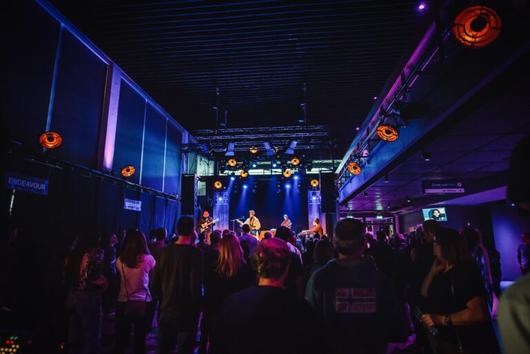 Een live optreden met muzikanten op een podium, omringd door een publiek in een sfeervolle, verlichte zaal.