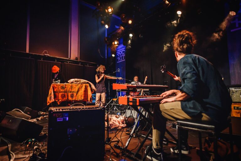 Een live muziekopvoering met verschillende artiesten op een podium, met instrumenten zoals een keyboard en een microfoon.