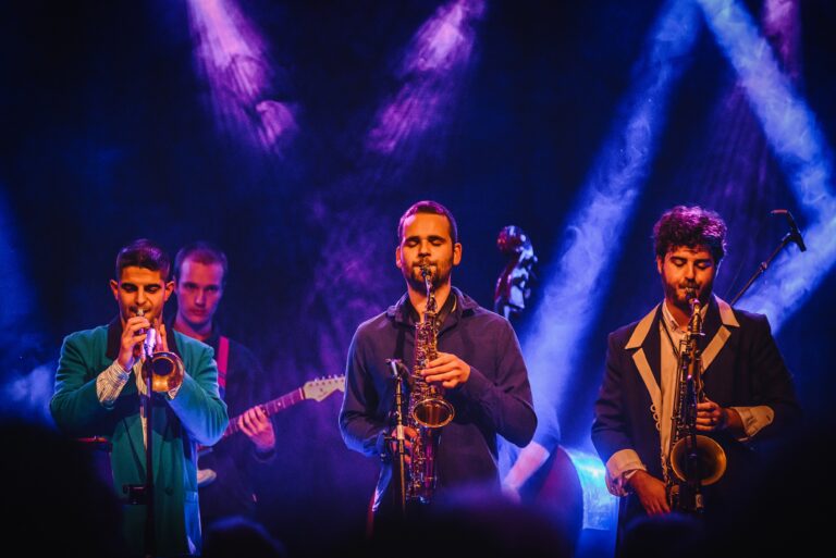 Een band speelt live muziek op een podium, met muzikanten die verschillende instrumenten bespelen onder kleurrijke lichteffecten.