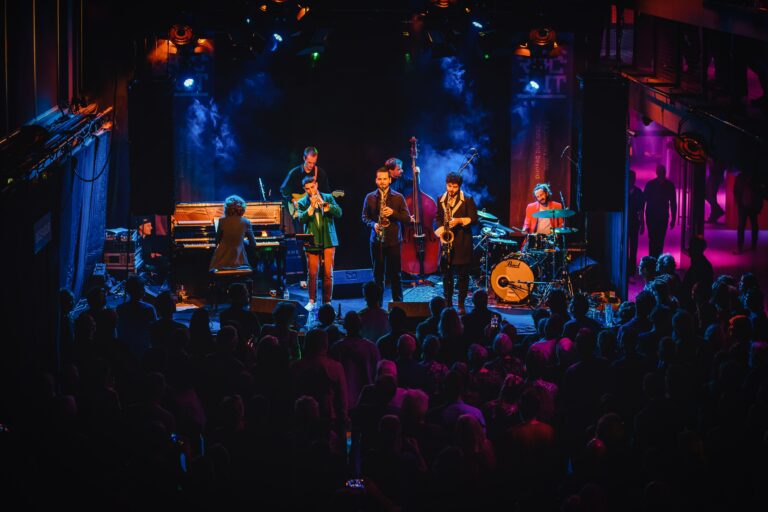 Een live band speelt op het podium voor een enthousiast publiek, omgeven door kleurrijke verlichting.