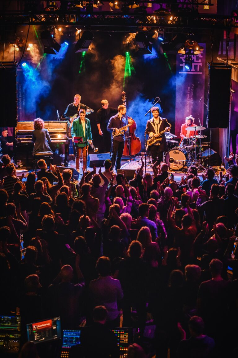 Een muziekgroep speelt op het podium voor een enthousiaste menigte, verlicht door kleurrijke lichten.