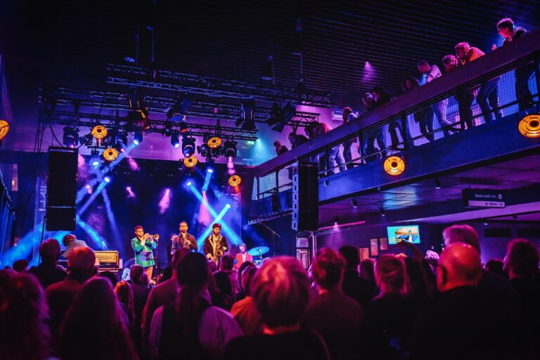 Een live muziekoptreden met een drukke menigte, felgekleurde lichten en een podium met artiesten.
