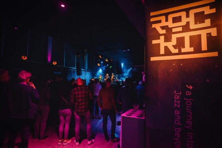 Een drukke muziekevenement locatie met een publiek dat naar een podium met blauwe en paarse verlichting kijkt.