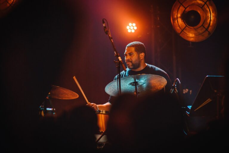 Een drummer speelt op een podium, verlicht door warme lichten in een donkere omgeving.