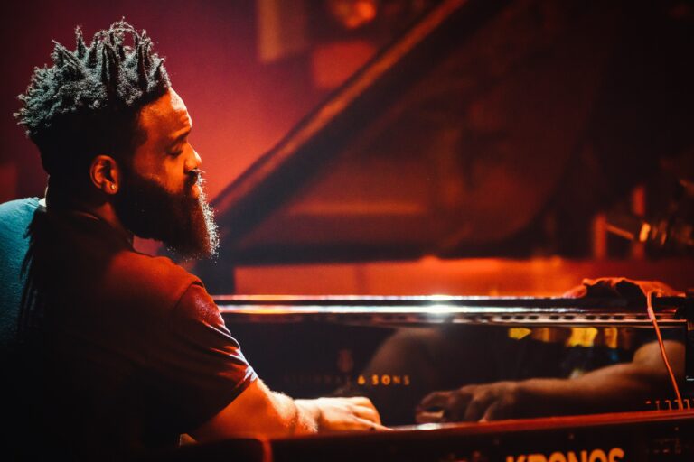 Een man met een baard speelt piano in een sfeervolle, verlichte omgeving.