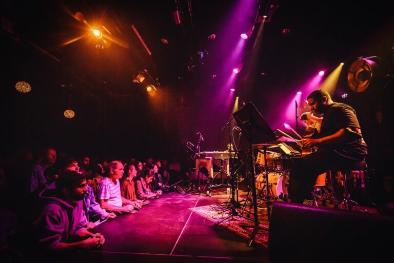 Een muzikant speelt op een podium met kleurrijke lichten, terwijl het publiek aandachtig kijkt.