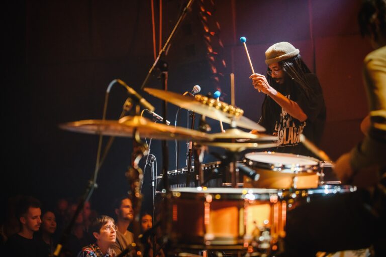 Een drummer speelt op een drumstel terwijl het publiek toekijkt in een sfeervolle omgeving.