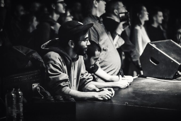 Een groep mensen kijkt aandachtig naar een evenement, met focus op twee mannen die aan een tafel zitten.