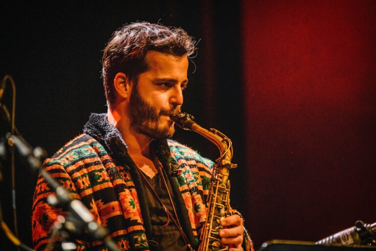 Een muzikant speelt saxophone, gekleed in een kleurrijke jas met een patroon, tegen een sfeervolle achtergrond.