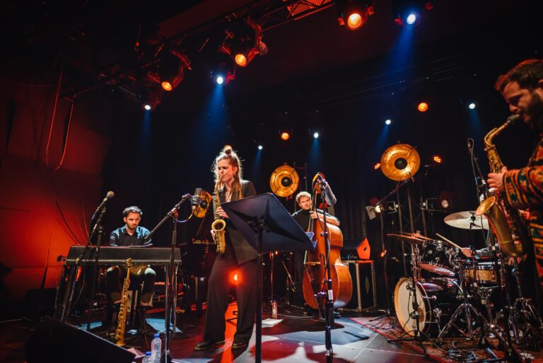 Een muzikant speelt saxofoon op een podium met andere bandleden, omringd door kleurrijke lichten.