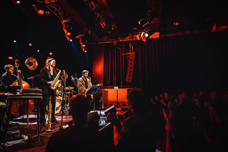 Een live muzieksessie met muzikanten op het podium, verlicht door warme lichten, terwijl het publiek toekijkt.