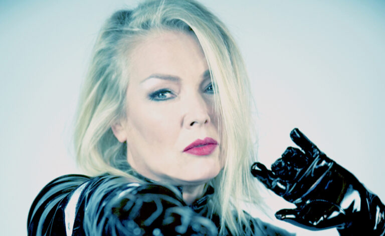 Portretfoto van Kim Wilde voor een witte muur met een zwart leren jasje aan.