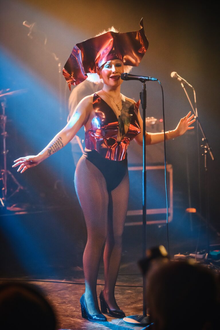 Een performer met een opvallend kostuum zingt op het podium, omringd door lichtstralen en instrumenten.