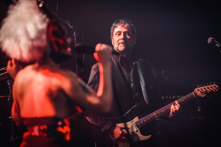 Een man met een gitaar speelt op een podium terwijl een zangeres voor hem zingt.