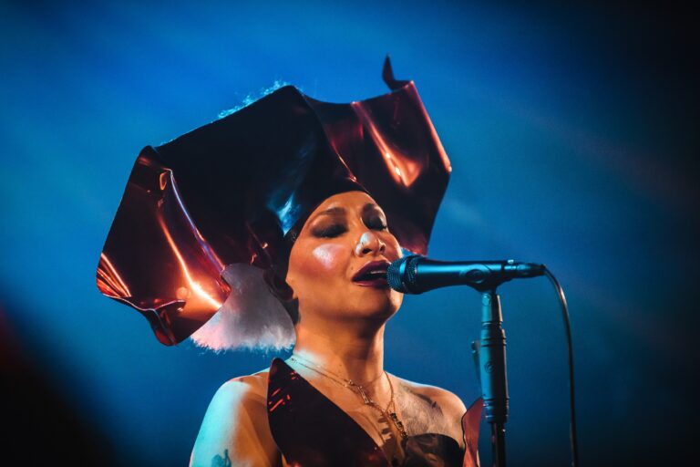 Een zangeres met een opvallende hoed en microfoon, die op het podium zingt met blauwe verlichting op de achtergrond.