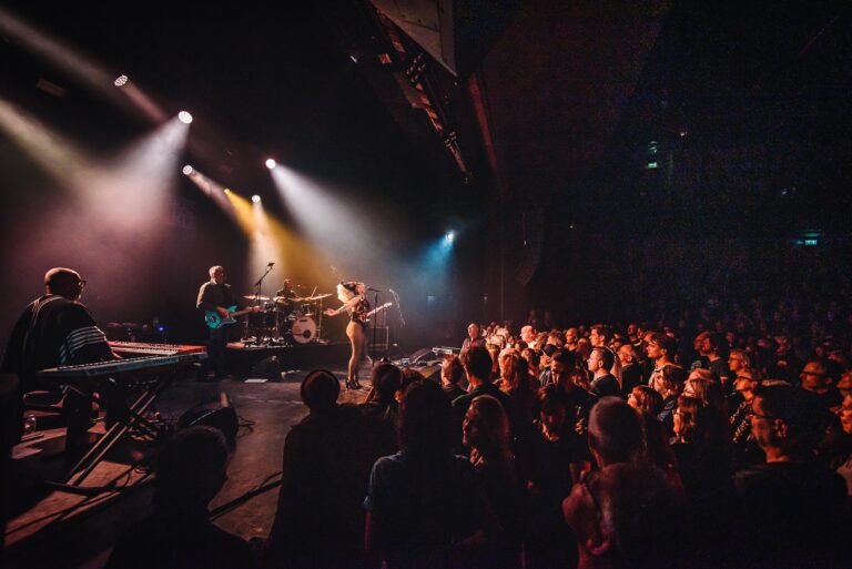 Een muzikant treedt op voor een enthousiast publiek in een donker concertzaaltje met kleurrijke lichten.