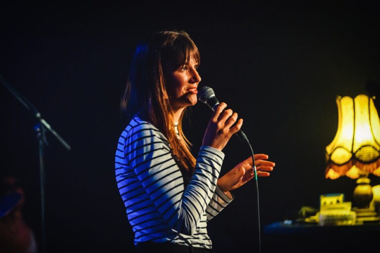 Een vrouw met een microfoon spreekt tijdens een optreden in een sfeervolle omgeving.