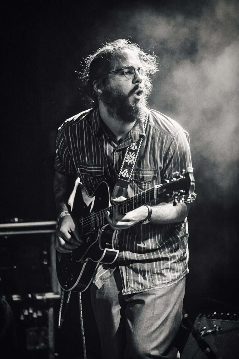 Een man speelt een elektrische gitaar op een podium, omringd door rook en in zwart-wit.