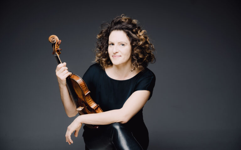 Een vrouw met krullend haar poseert met een hobo in een zwarte outfit tegen een neutrale achtergrond.